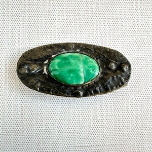 May include: Oval-shaped antique brooch with a bright green stone center. The stone is set in a dark, textured metal frame with decorative detailing. A vintage jewelry piece.