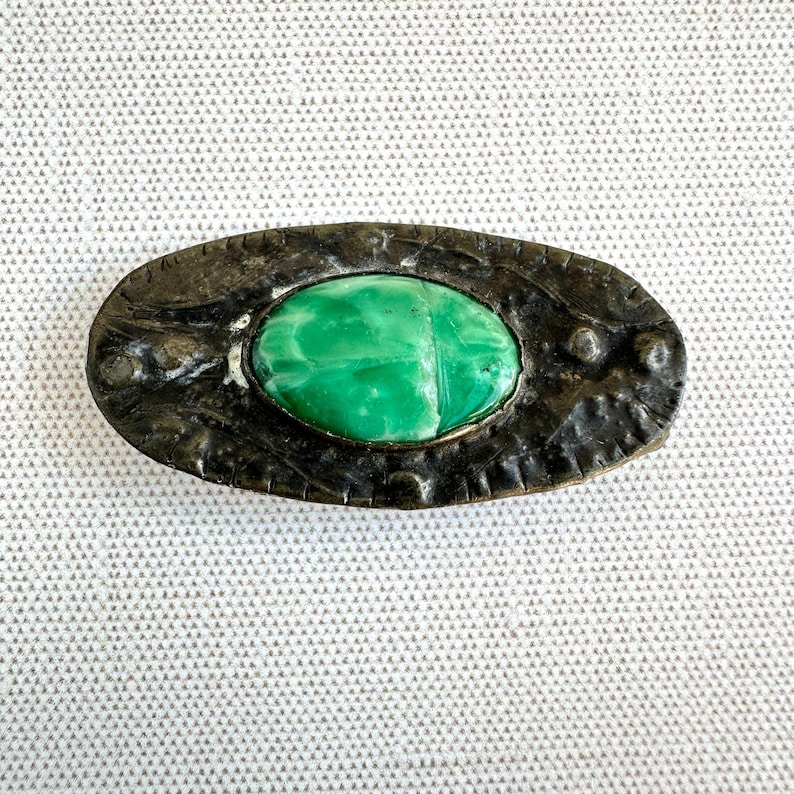 May include: Oval-shaped antique brooch with a bright green stone center. The stone is set in a dark, textured metal frame with decorative detailing. A vintage jewelry piece.