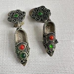 May include: A pair of silver-toned earrings featuring a floral design with green gemstones. Each earring has a shoe-shaped pendant with red and green gemstones. The earrings are set against a white textured background.