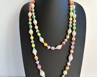 May include: A colorful beaded necklace with a variety of round beads in shades of red, orange, yellow, green, and pink. The beads are interspersed with white, openwork sections and small, clear star-shaped accents. The necklace is displayed on a black mannequin.