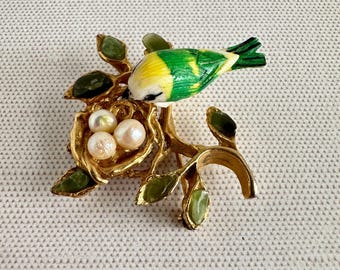 May include: A gold-toned bird brooch featuring a green and yellow bird perched on a branch with a nest. The nest contains three pearls, and the branch has green leaf accents. A unique piece of jewelry.