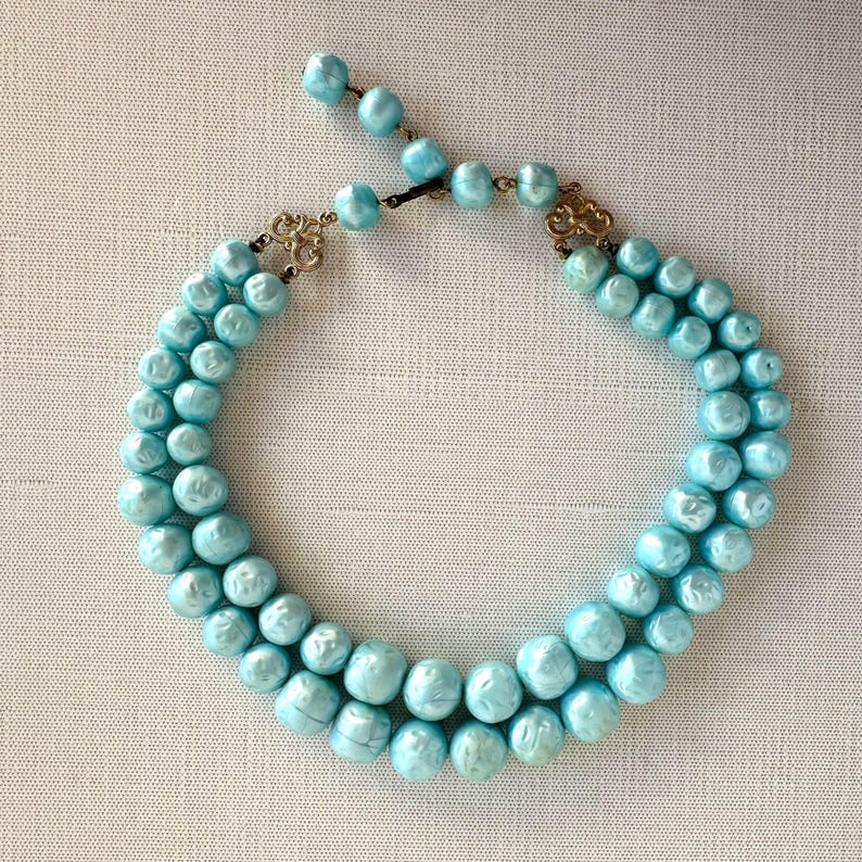 May include: A vintage, light blue, double-strand beaded necklace. The necklace features round, faux pearls in a soft, pastel blue hue. The clasp and connecting hardware are gold-toned. The necklace is displayed on a neutral, textured surface.
