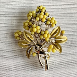 May include: A vintage brooch in the shape of a floral bouquet. The brooch features clusters of yellow beads, gold-toned leaves, and a ribbon detail. The design is intricate and detailed, with a classic, timeless appeal.