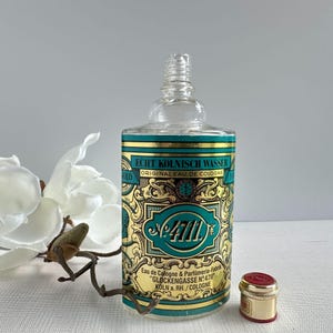 May include: A clear glass cologne bottle with a decorative label featuring gold and teal accents. The label reads "ECHT K&Ouml;LNISCH WASSER ORIGINAL EAU DE COLOGNE" and "No. 4711". A small, gold-colored cap with a red top sits beside the bottle.