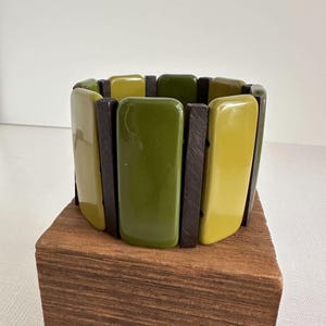 Könnte beinhalten: Ein breites Armband mit abwechselnden rechteckigen Segmenten in Olivgrün, Gelbgrün und Dunkelbraun. Das Armband besteht aus Holz und Harz und wird auf einem Holzblock präsentiert.