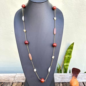 Pode incluir: Um colar longo com contas alternadas em vermelho escuro, castanho claro e branco. As contas são conectadas por uma delicada corrente dourada. O colar é exibido em um manequim preto.