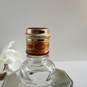 May include: Close-up of a clear glass bottle with a decorative cap. The cap is silver and red with gold accents and the text "47th". The bottle has a geometric shape.