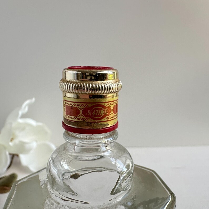 May include: Close-up of a clear glass bottle with a decorative cap. The cap is silver and red with gold accents and the text "47th". The bottle has a geometric shape.