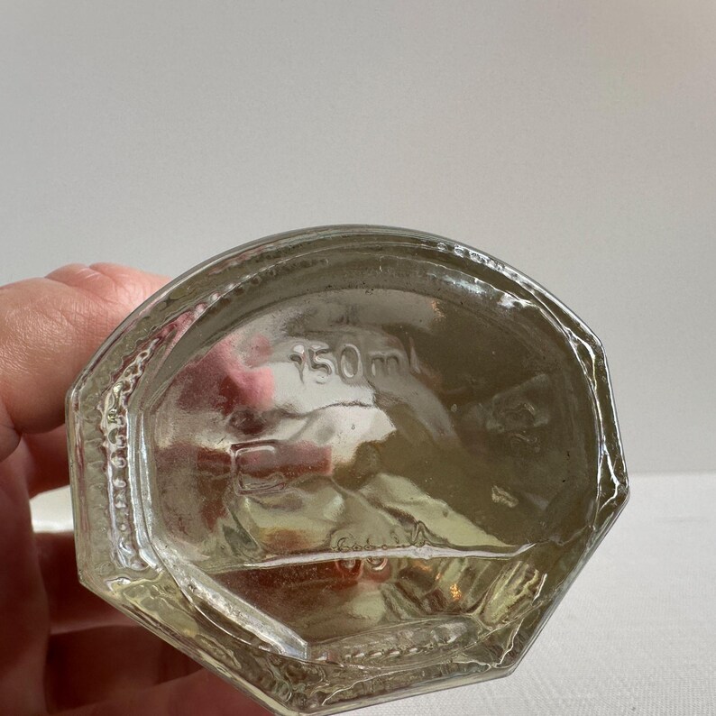 May include: Clear glass bottle with a geometric design. The bottom of the bottle has the text "150 ml". The bottle is partially filled with a light-colored liquid.