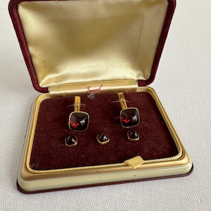 Puede incluir: Un juego de gemelos dorados y tres botones a juego con piedras facetadas de color rojo oscuro cuadrado. Los artículos se presentan en una caja con bisagras con una tapa interior de color crema marcada con la marca Swank y una base de terciopelo granate.