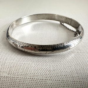以下が含まれることがあります： 彫刻された花柄と幾何学模様が施されたシルバーのバングルブレスレット。ヒンジ付きブレスレットには安全チェーンが付いています。クラシックなジュエリーです。
