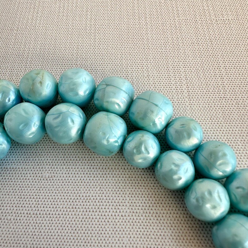 May include: Close-up of a strand of light blue, round beads. The beads have a textured surface and a pearlescent sheen. The beads are arranged in a curved line against a light gray background.