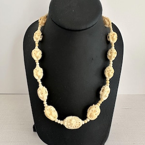 May include: Ivory-colored necklace with carved floral and oval beads. The necklace is displayed on a black mannequin. The beads have intricate details and are strung together with a matching cord. The necklace is a vintage piece.