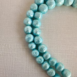 May include: A close-up of a necklace featuring large, light blue beads. The beads have a textured, irregular surface and are strung together. The necklace is displayed on a light gray textured surface.