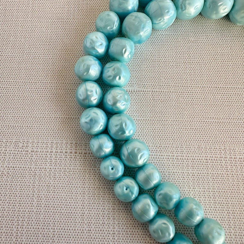 May include: A close-up of a necklace featuring large, light blue beads. The beads have a textured, irregular surface and are strung together. The necklace is displayed on a light gray textured surface.