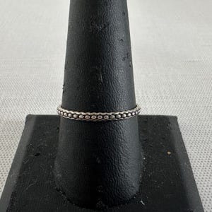 Puede incluir: Un anillo de plata con un diseño de cuentas. El anillo se muestra sobre un soporte de joyería negro sobre un fondo blanco. La banda del anillo está adornada con pequeñas cuentas espaciadas uniformemente, creando una apariencia texturizada. El anillo es una joya sencilla y elegante.
