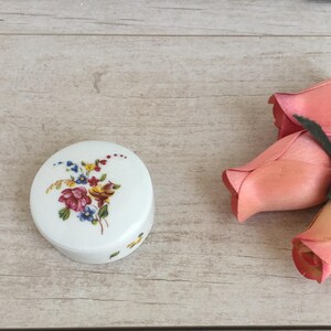 May include: A small, white porcelain trinket box with a floral design featuring red, yellow, and blue flowers. The box is round and has a lid.