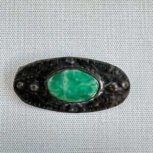 May include: Oval-shaped, antique-style brooch with a dark, textured metal frame. The brooch features a vibrant, marbled green stone center. The frame has small, raised circular details around the edge, adding to its vintage aesthetic.