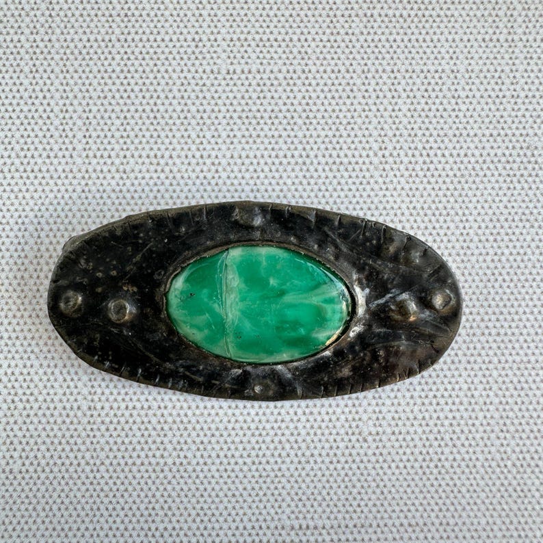 May include: Oval-shaped, antique-style brooch with a dark, textured metal frame. The brooch features a vibrant, marbled green stone center. The frame has small, raised circular details around the edge, adding to its vintage aesthetic.