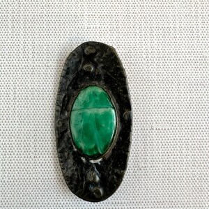 May include: An oval-shaped, dark metal pendant with a carved green scarab beetle at its center. The pendant has a textured border and small decorative accents. The beetle is a vibrant green color.