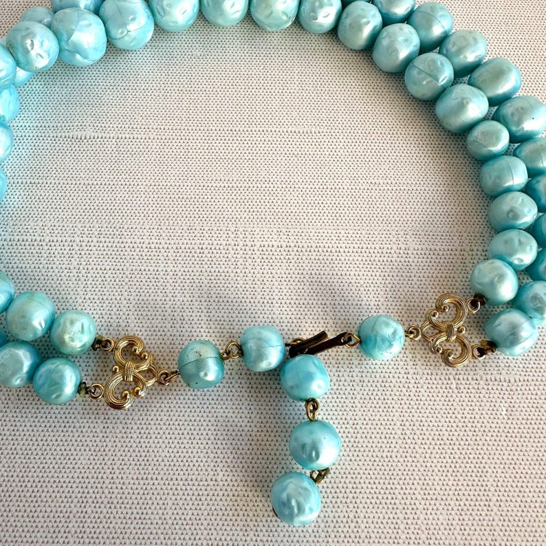 May include: A vintage necklace featuring two strands of light blue, pearlescent beads. The necklace has ornate gold-tone clasps and a dangling strand of beads. The beads are round and have a slightly irregular shape.