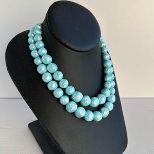 May include: A two-strand necklace featuring large, round, light blue beads. The beads have a pearlescent sheen and are displayed on a black jewelry bust. The necklace is a statement piece.