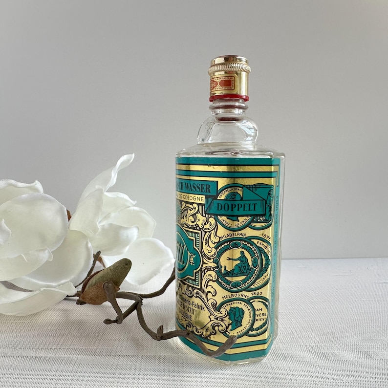 May include: A vintage cologne bottle with a gold and teal label featuring the text "SCH WASSER COLOGNE DOPPELT". The bottle has a gold cap with red accents. A white flower and a decorative metal piece are in the foreground.