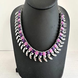 Puede incluir: Un collar vintage con una cadena y detalles de metal plateado. El collar presenta secciones alternas de incrustaciones rectangulares de esmalte morado y rosa. El diseño incluye elementos metálicos curvos en forma de garra.