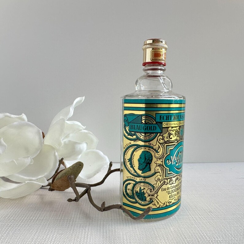 May include: A vintage glass cologne bottle with a gold and teal label featuring the text "BLAU GOLD" and "Echt K&ouml;lnisch Wasser". The bottle has a gold cap and a red band. A decorative metal branch and white flower are in the foreground.