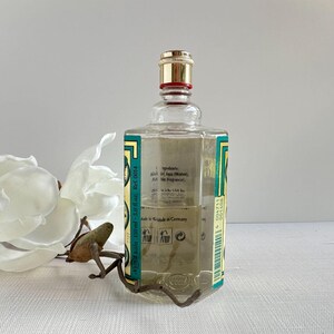 May include: A clear glass cologne bottle with a gold cap and a red band. The bottle has a green and gold label with text and is 5.0 fl oz. The cologne is in a clear glass bottle with a decorative metal stand.