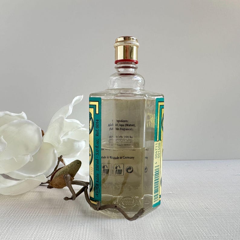 May include: A clear glass cologne bottle with a gold cap and a red band. The bottle has a green and gold label with text and is 5.0 fl oz. The cologne is in a clear glass bottle with a decorative metal stand.