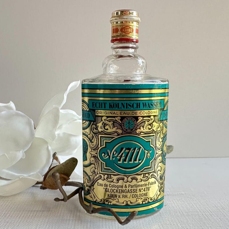 May include: A vintage glass cologne bottle with a gold and teal label. The label reads "ECHT KOLNISCH WASSER ORIGINAL EAU DE COLOGNE" and "No. 4711". The bottle has a gold cap with red accents.