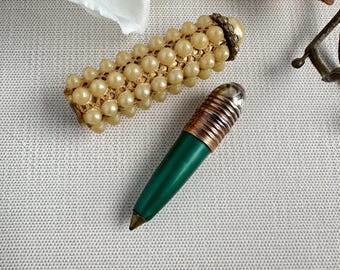 May include: Two vintage writing instruments. One is a gold-toned lipstick-shaped pen covered in faux pearls. The other is a green pen with a silver and copper-toned cap. Both are on a textured white surface.