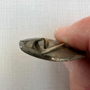 May include: Close-up of a vintage metal pin with a curved clasp and a sharp pin. The pin has a tarnished, aged appearance, with visible wear and discoloration. The clasp is a contrasting brass color.