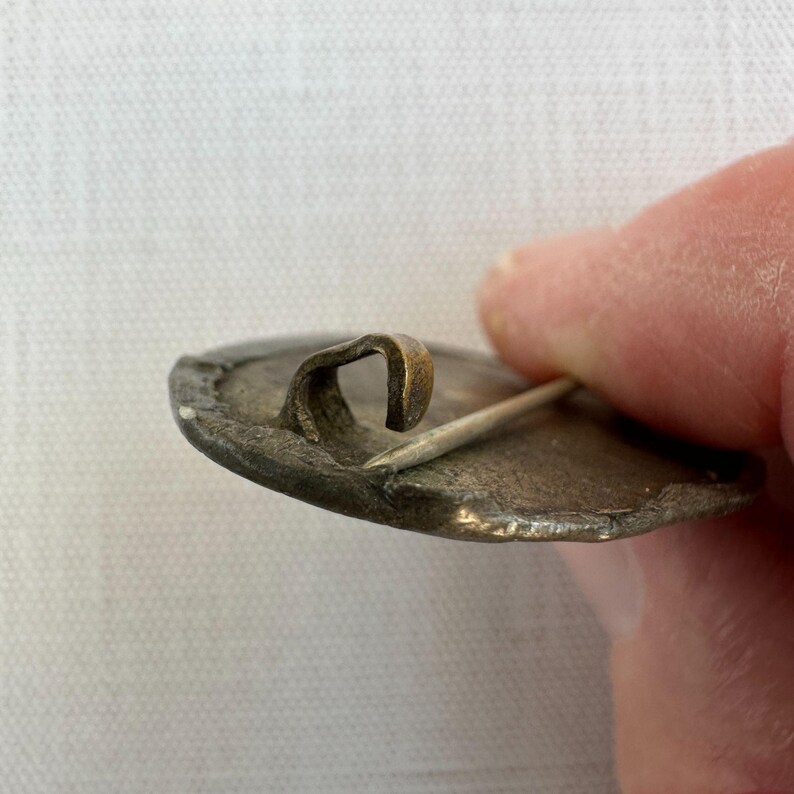 May include: Close-up of a vintage metal pin with a curved clasp and a sharp pin. The pin has a tarnished, aged appearance, with visible wear and discoloration. The clasp is a contrasting brass color.