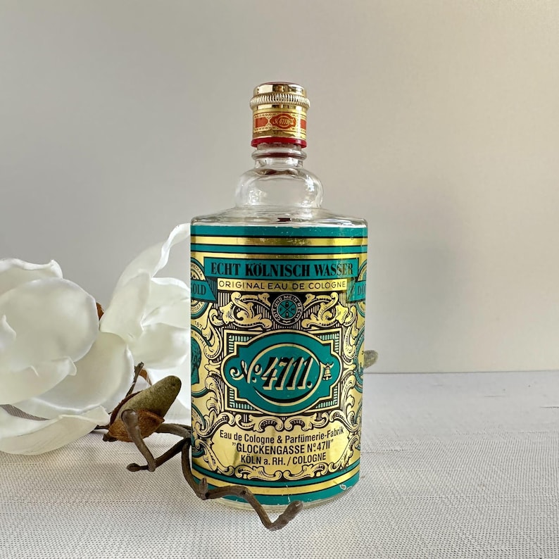 May include: A vintage glass cologne bottle with a gold and teal label. The label reads "ECHT K&Ouml;LNISCH WASSER ORIGINAL EAU DE COLOGNE" and "4711". The bottle has a gold cap and is displayed with a white flower.