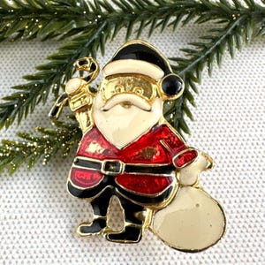 May include: A gold-toned Santa Claus brooch with a black hat and boots, red suit, and white beard. The brooch holds a candy cane and a beige sack. The brooch is set against a backdrop of green pine needles.