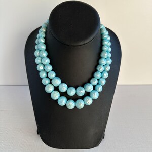 May include: A two-strand necklace featuring large, round, light blue beads. The necklace is displayed on a black mannequin bust against a white background. The beads have a pearlescent sheen.