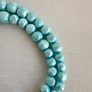 May include: A necklace featuring two strands of light blue, round beads. The beads have a slightly irregular shape and a pearlescent finish, creating a subtle shimmer. The necklace is displayed against a neutral background.