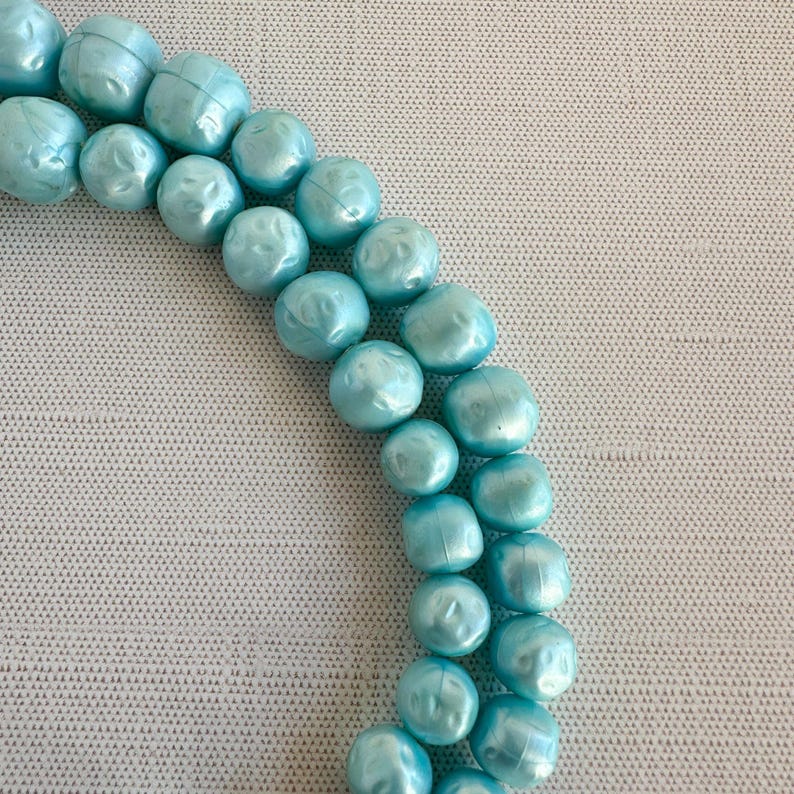 May include: A necklace featuring two strands of light blue, round beads. The beads have a slightly irregular shape and a pearlescent finish, creating a subtle shimmer. The necklace is displayed against a neutral background.