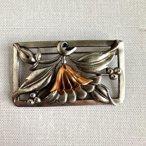 May include: A rectangular silver-toned brooch with a floral design. The brooch features a central flower with gold-colored petals, surrounded by silver leaves and berries. The frame has a simple, geometric design.
