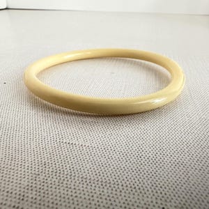 May include: A cream-colored, round bangle bracelet. The smooth, polished surface reflects light, highlighting its simple, elegant design. This classic accessory is a versatile addition to any jewelry collection.