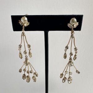 May include: A pair of vintage gold-toned chandelier earrings. Each earring features a cluster of clear, sparkling rhinestones at the top, with delicate, branching strands adorned with more rhinestones. The earrings are displayed on a black stand against a white background.