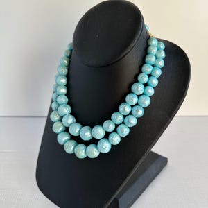 May include: A two-strand necklace featuring large, round, light blue beads. The necklace is displayed on a black jewelry stand. The beads have a pearlescent sheen, and the necklace is fastened with a gold-colored clasp.