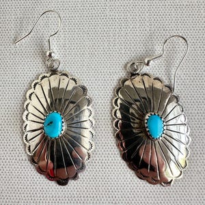May include: A pair of silver-toned earrings with a turquoise stone center. The earrings have a sunburst design with a scalloped edge and a hook closure. The earrings are oval-shaped and are displayed on a textured surface.