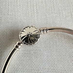 May include: A silver bracelet featuring a floral clasp. The clasp has a detailed, textured design with a small star-like element in the centre. The bracelet's metal has a polished, reflective surface.
