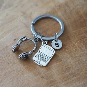 May include: Silver-toned keychain with charms of headphones, a laptop, and a circular tag with the letter "S". The keychain rests on a wooden surface. The headphones and laptop charms are detailed.