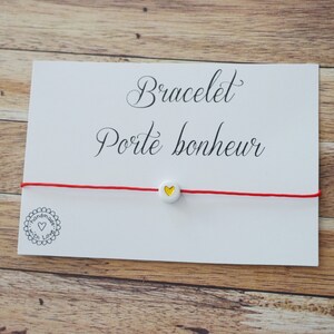 Peut inclure: Un bracelet rouge avec une perle blanche ornée d'un cœur jaune. Le bracelet est présenté sur une carte blanche avec les mots "Bracelet Porte bonheur" en écriture élégante. La carte comporte également un petit logo en forme de cœur.