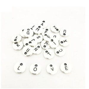 May include: A collection of silver metal alphabet letter charms with black lettering. The charms are round and have two small holes at the top.
