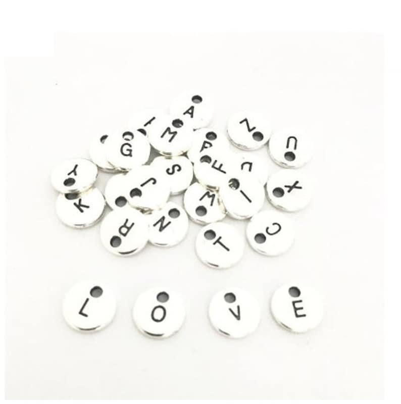May include: A collection of silver metal alphabet letter charms with black lettering. The charms are round and have two small holes at the top.
